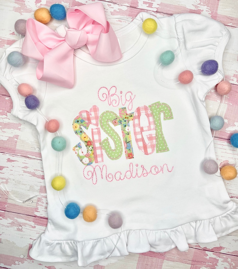 May include: White t-shirt with a pink bow on top. The shirt has a green and pink patchwork design that says "Big Sister" with the name "Madison" embroidered underneath.