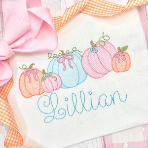 Pu&ograve; includere: Una camicia bianca ricamata con una fila di cinque zucche color pastello e il nome "Lillian" ricamato in filo blu.