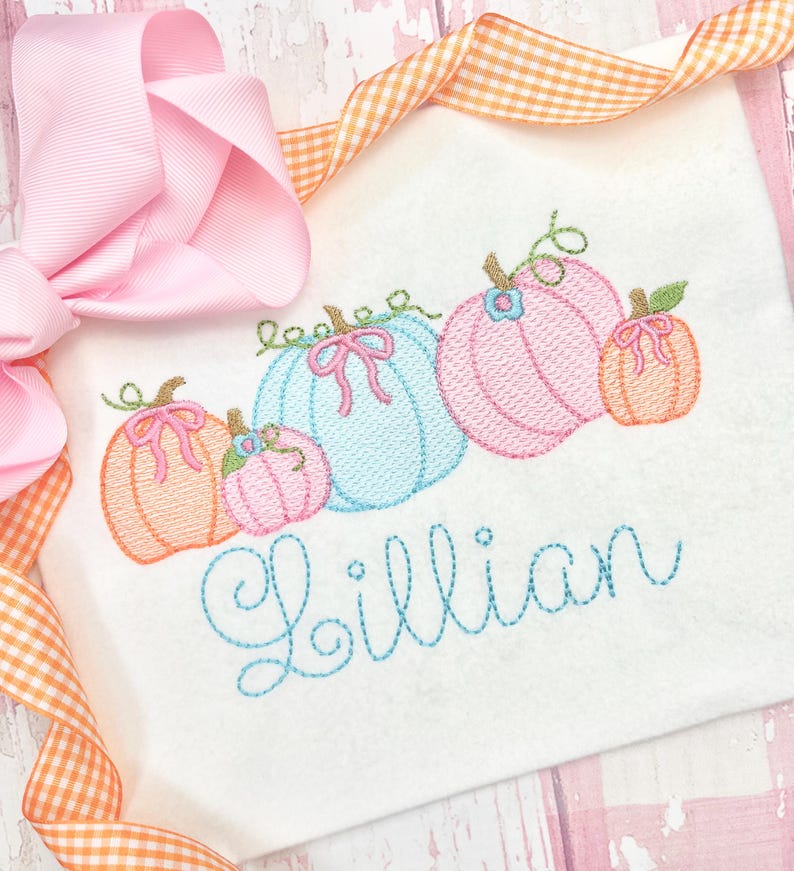 Pu&ograve; includere: Una camicia bianca ricamata con una fila di cinque zucche color pastello e il nome "Lillian" ricamato in filo blu.