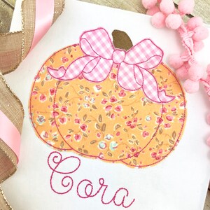 May include: A white fabric with a pink and orange pumpkin with a pink gingham bow and the name "Cora" embroidered in pink thread.