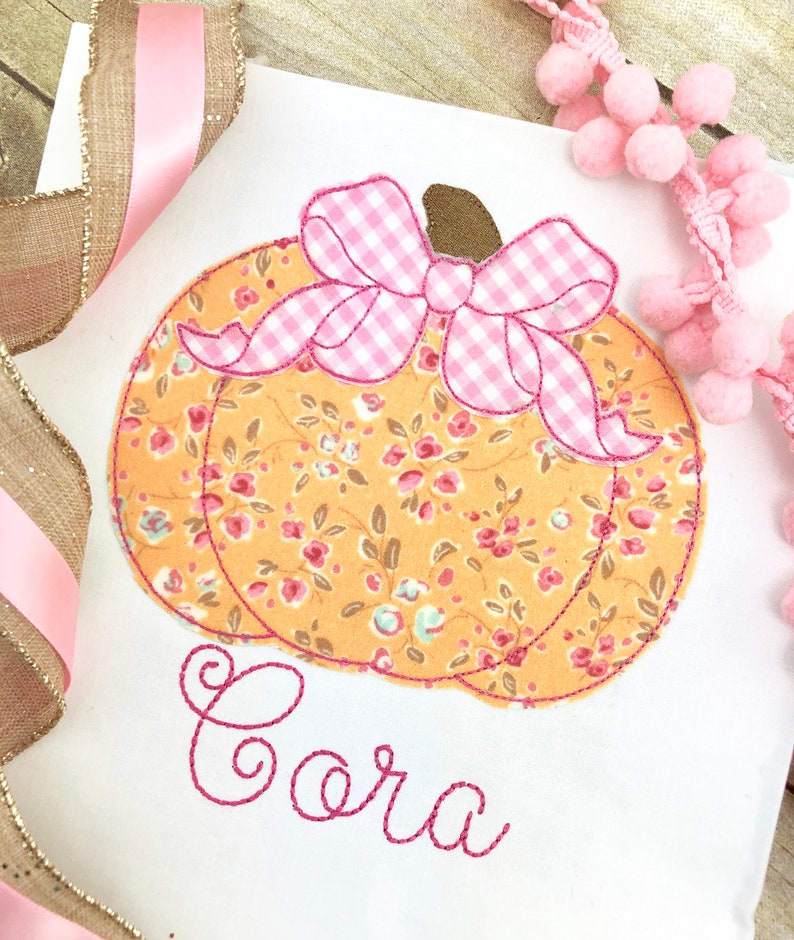 May include: A white fabric with a pink and orange pumpkin with a pink gingham bow and the name "Cora" embroidered in pink thread.
