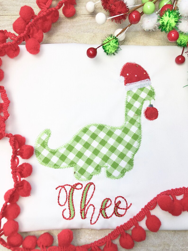 May include: A white onesie with a green and white gingham dinosaur wearing a red Santa hat and holding a red ornament. The name "Theo" is embroidered in green and red on the onesie.