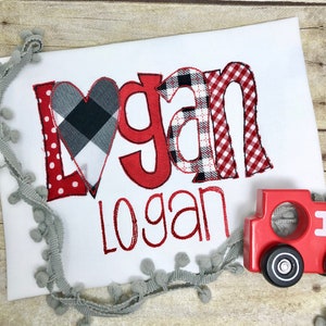 May include: A white babygrow with the name "Logan" embroidered in red and black checked fabric. The name is in a large, playful font with a red heart in the middle of the "L".