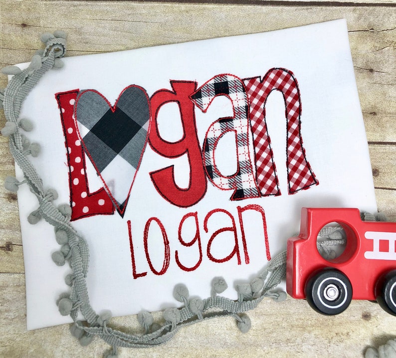 May include: A white babygrow with the name "Logan" embroidered in red and black checked fabric. The name is in a large, playful font with a red heart in the middle of the "L".