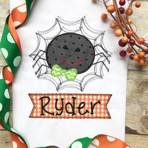 May include: Embroidered design of a black spider with a green bow tie on a white background. The spider is in a web and the name "Ryder" is embroidered in orange and white checkered fabric below the spider.