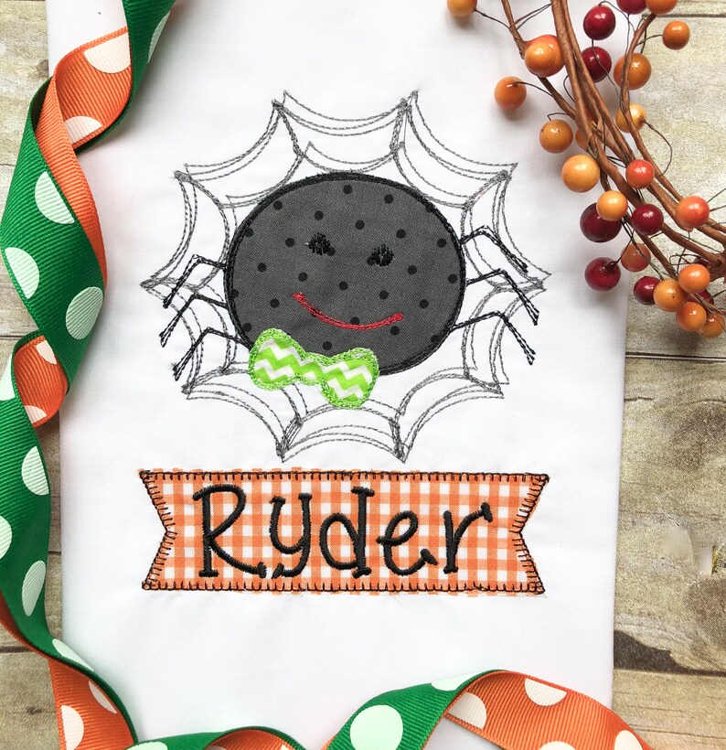 May include: Embroidered design of a black spider with a green bow tie on a white background. The spider is in a web and the name "Ryder" is embroidered in orange and white checkered fabric below the spider.