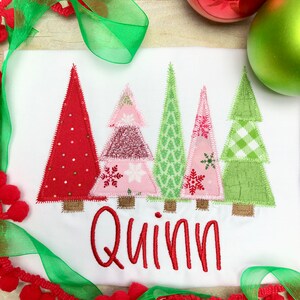Puede incluir: Un pelele blanco con un nombre bordado en rojo "Quinn" y cuatro &aacute;rboles de Navidad de patchwork coloridos. Los &aacute;rboles son rojo, rosa, verde y verde con detalles blancos.