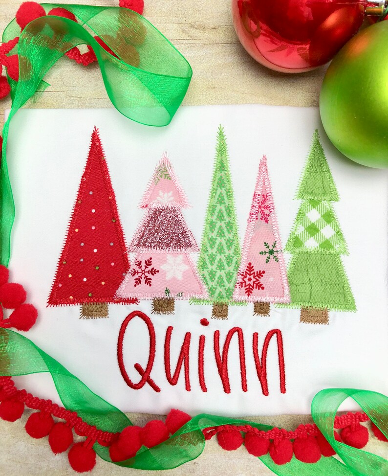 Puede incluir: Un pelele blanco con un nombre bordado en rojo "Quinn" y cuatro &aacute;rboles de Navidad de patchwork coloridos. Los &aacute;rboles son rojo, rosa, verde y verde con detalles blancos.