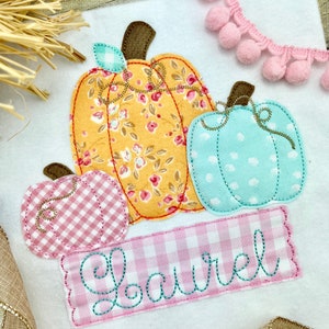 May include: A pink, blue, and yellow fabric applique of three pumpkins with the name "Laurel" stitched in a pink and green banner.