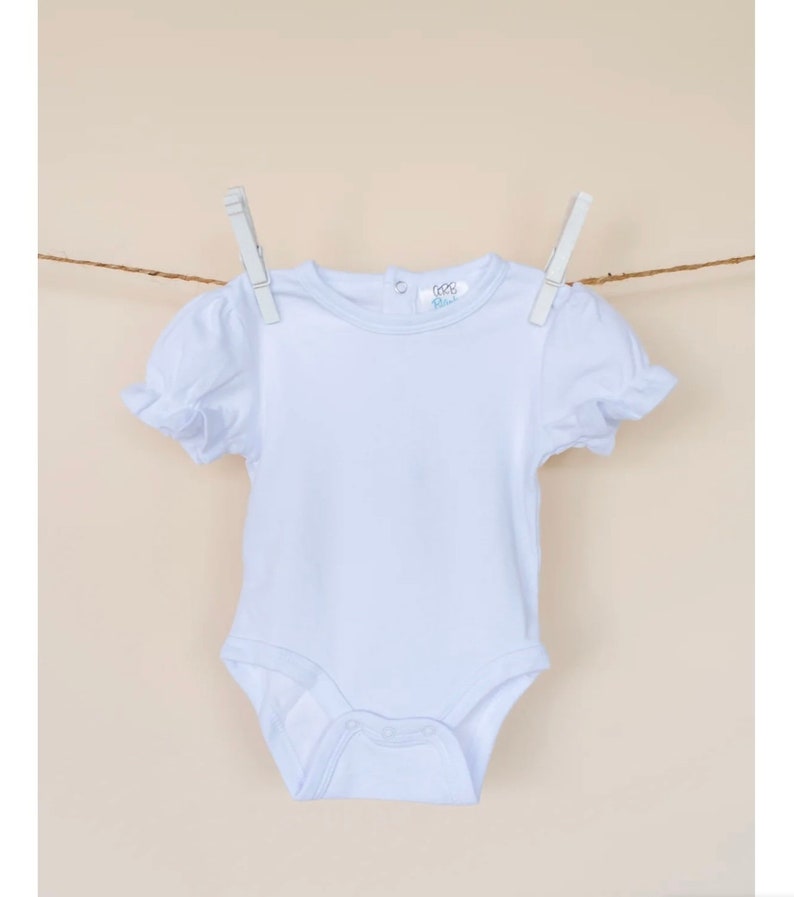 May include: White baby onesie with short sleeves and a ruffled trim. The onesie has a round neckline and a snap closure at the bottom.