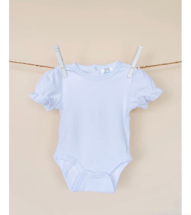 Puede incluir: Un body blanco para beb&eacute; con mangas cortas y un borde con volantes. El body tiene un escote redondo y un cierre de botones a presi&oacute;n en la parte inferior.