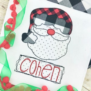 May include: White fabric with a Santa Claus face design. The Santa face features a red and black buffalo plaid hat, a black and white beard, and a red nose. The name "Cohen" is embroidered in red.
