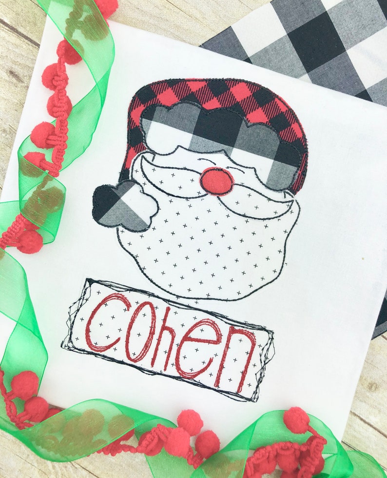 May include: White fabric with a Santa Claus face design. The Santa face features a red and black buffalo plaid hat, a black and white beard, and a red nose. The name "Cohen" is embroidered in red.