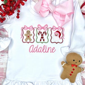 May include: White long-sleeved shirt with a ruffle hem, featuring embroidered Christmas designs: a gingerbread man, candy canes, and Santa Claus. The name "Adaline" is embroidered in pink. A large pink bow is at the top.