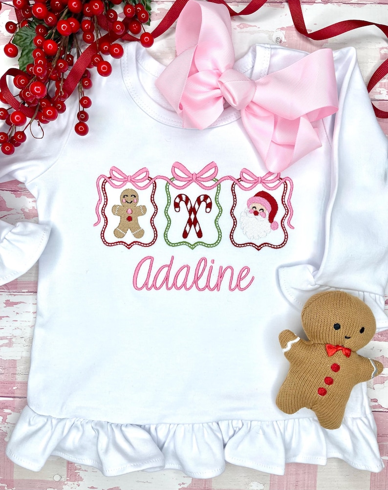 May include: White long-sleeved shirt with a ruffle hem, featuring embroidered Christmas designs: a gingerbread man, candy canes, and Santa Claus. The name "Adaline" is embroidered in pink. A large pink bow is at the top.