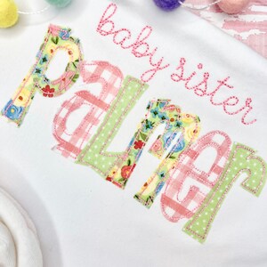 May include: White baby onesie with pink and green embroidered letters that spell "baby sister Palmer".