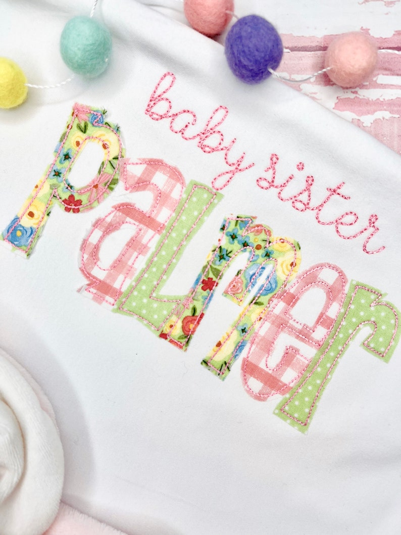 May include: White baby onesie with pink and green embroidered letters that spell "baby sister Palmer".