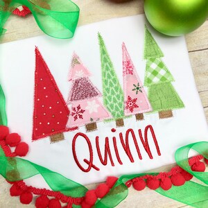 Puede incluir: Un pelele blanco con un dise&ntilde;o de patchwork rojo, rosa y verde de cinco &aacute;rboles de Navidad. El pelele tiene el nombre "Quinn" bordado en rojo debajo de los &aacute;rboles.