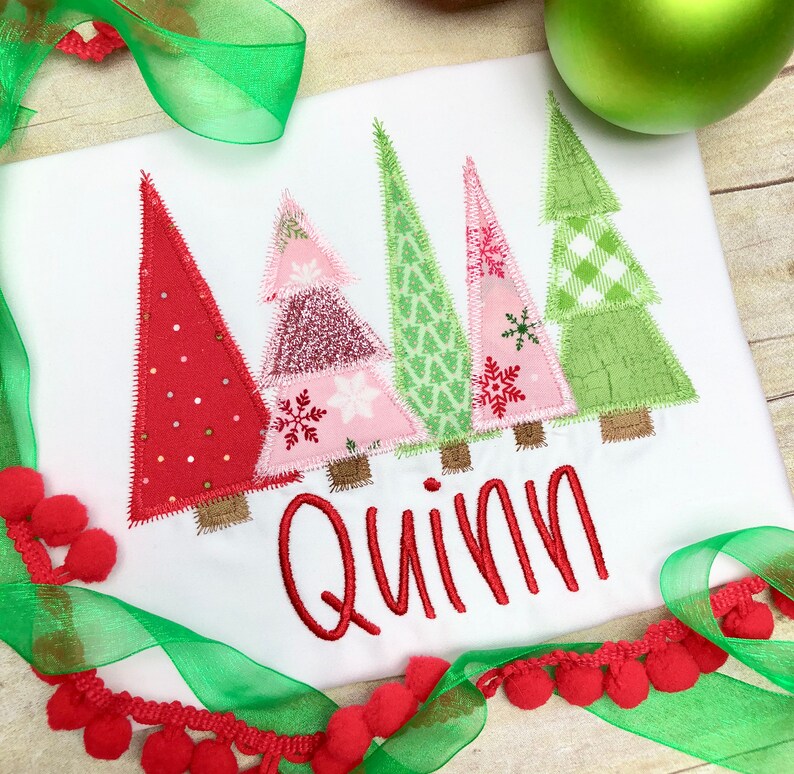 Puede incluir: Un pelele blanco con un dise&ntilde;o de patchwork rojo, rosa y verde de cinco &aacute;rboles de Navidad. El pelele tiene el nombre "Quinn" bordado en rojo debajo de los &aacute;rboles.