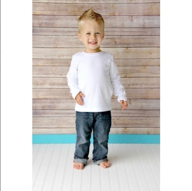 May include: A young boy wearing a white long-sleeved shirt and blue jeans stands in front of a wood panel background.