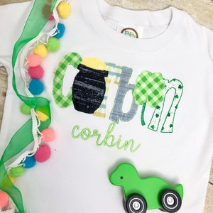 Op de afbeelding: Een wit shirt met lange mouwen met een groen, blauw en geel geborduurd ontwerp dat "Corbin" zegt met een pot goud en klavertjes. Een groene speelgoedtruck bevindt zich op het shirt.