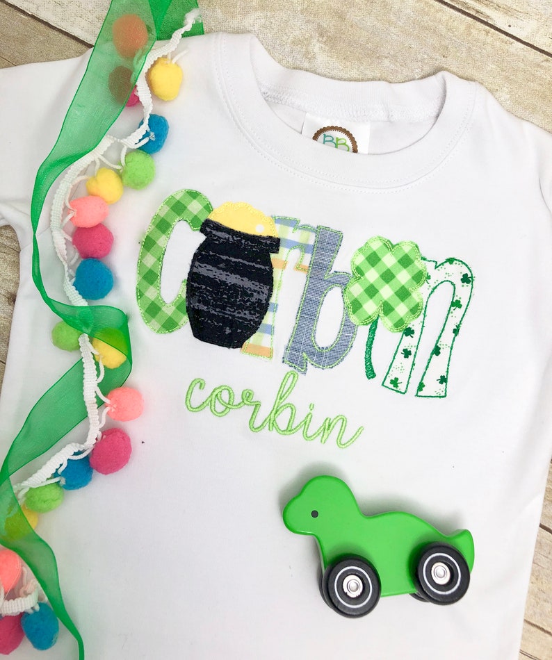 Op de afbeelding: Een wit shirt met lange mouwen met een groen, blauw en geel geborduurd ontwerp dat "Corbin" zegt met een pot goud en klavertjes. Een groene speelgoedtruck bevindt zich op het shirt.