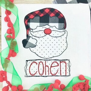 May include: White square with a Santa Claus design. The Santa's hat is red and black buffalo plaid, with a matching cuff. The beard is white with small black crosses. The name "COHEN" is in red.