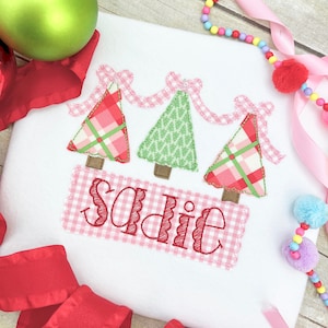 May include: White Christmas-themed shirt with three pink and green trees with pink bows and a pink gingham banner with the name "Sadie" embroidered on it.