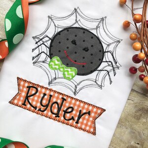 May include: A white t-shirt with an embroidered black spider wearing a green bow tie in a web. The shirt has an orange and white checkered banner with the name "Ryder" embroidered on it.
