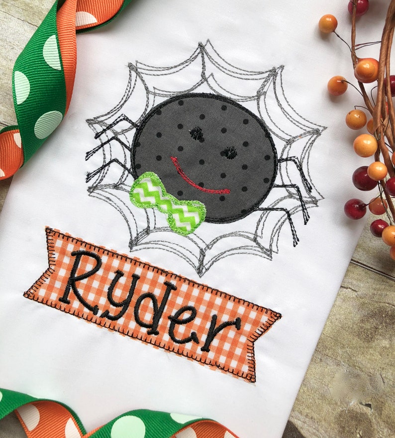 May include: A white t-shirt with an embroidered black spider wearing a green bow tie in a web. The shirt has an orange and white checkered banner with the name "Ryder" embroidered on it.