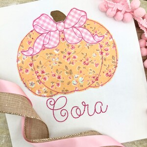 May include: A white fabric pumpkin with a pink and white gingham bow and floral print. The name "Cora" is embroidered in pink thread below the pumpkin.