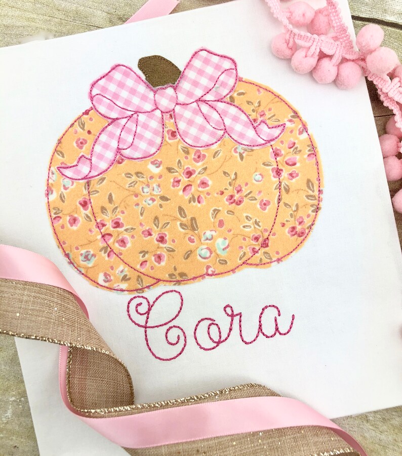 May include: A white fabric pumpkin with a pink and white gingham bow and floral print. The name "Cora" is embroidered in pink thread below the pumpkin.
