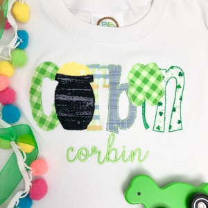 Op de afbeelding: Een witte baby romper met groene, gele en blauwe applicatie letters die "corbin" spellen met een pot goud en klavertjevier accenten. De romper ligt op een wit oppervlak met een groen speelgoed.