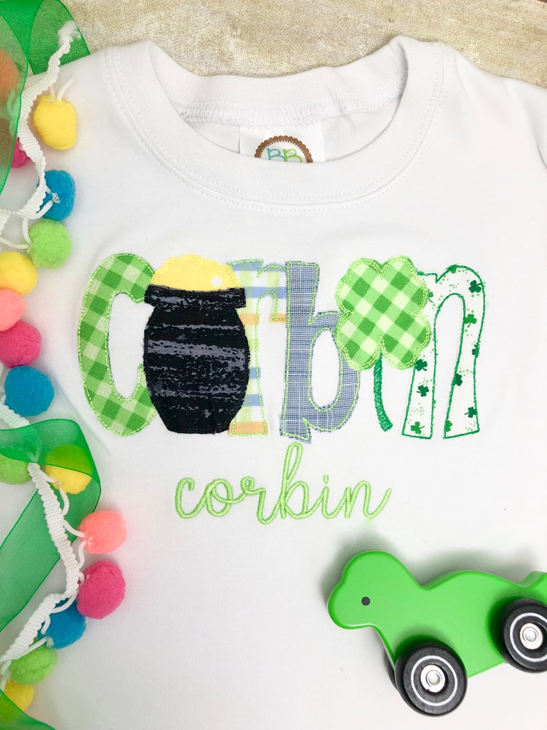 Op de afbeelding: Een witte baby romper met groene, gele en blauwe applicatie letters die "corbin" spellen met een pot goud en klavertjevier accenten. De romper ligt op een wit oppervlak met een groen speelgoed.