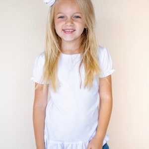 Puede incluir: Una ni&ntilde;a joven con el pelo largo y rubio que lleva una camisa blanca de manga corta con un ribete de volantes. Est&aacute; sonriendo y lleva un lazo blanco en el pelo.