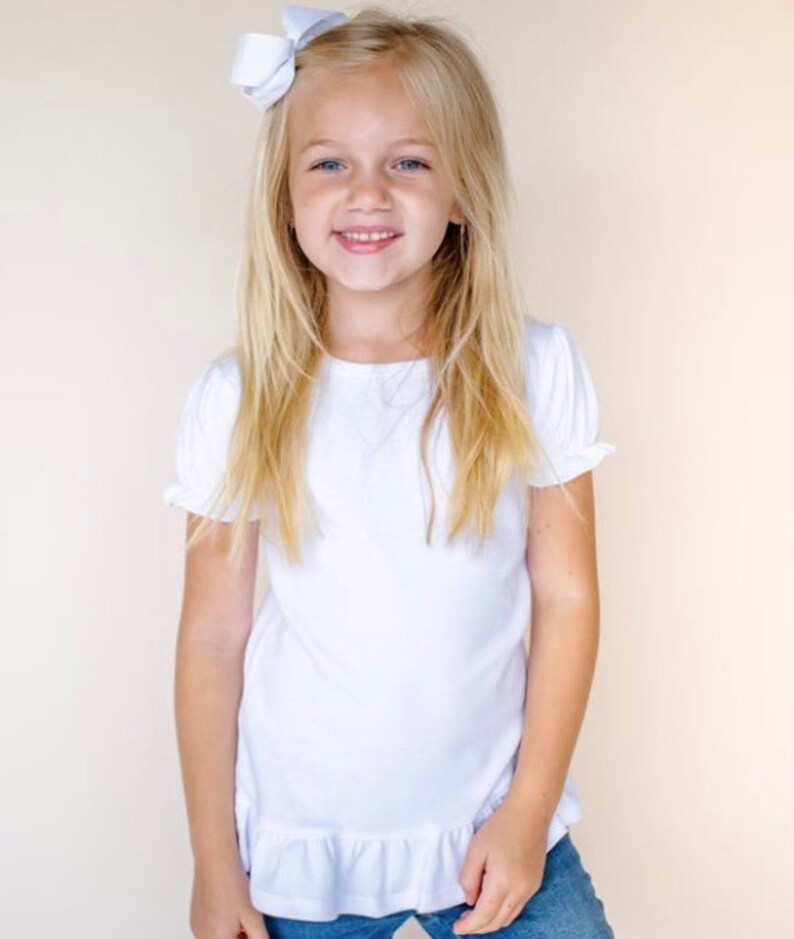 Puede incluir: Una ni&ntilde;a joven con el pelo largo y rubio que lleva una camisa blanca de manga corta con un ribete de volantes. Est&aacute; sonriendo y lleva un lazo blanco en el pelo.