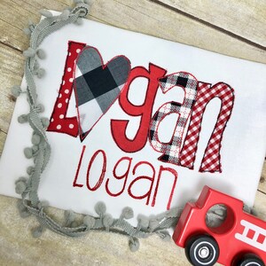 May include: A white onesie with the name "Logan" embroidered in red. The name is spelled out in large, red letters with a black and white checked heart in the middle. The onesie is next to a red toy fire engine.