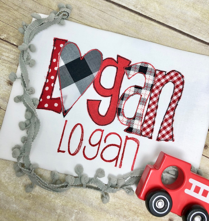 May include: A white onesie with the name "Logan" embroidered in red. The name is spelled out in large, red letters with a black and white checked heart in the middle. The onesie is next to a red toy fire engine.