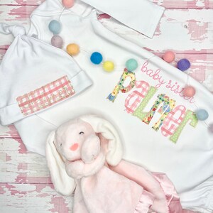 May include: A white baby gown with long sleeves and a pink and green gingham pattern. The gown has the text "baby sister Palmer" embroidered on the front. A white hat with a pink and white gingham pattern is included. A pink bunny lovey is also included.