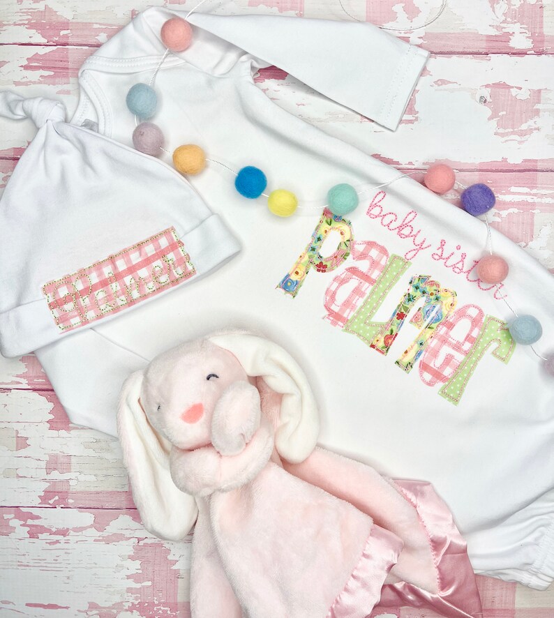 May include: A white baby gown with long sleeves and a pink and green gingham pattern. The gown has the text "baby sister Palmer" embroidered on the front. A white hat with a pink and white gingham pattern is included. A pink bunny lovey is also included.