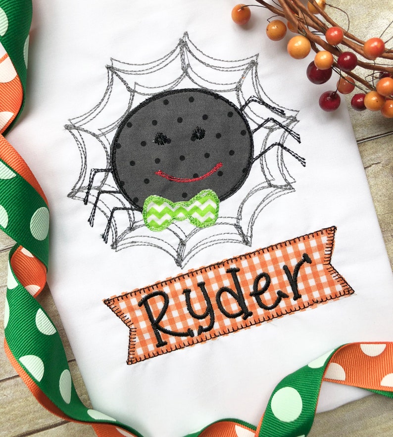 May include: A white onesie with a black spider wearing a green bow tie in a gray spider web. The onesie has a banner with the name "Ryder" in black text on an orange and white checkered background.