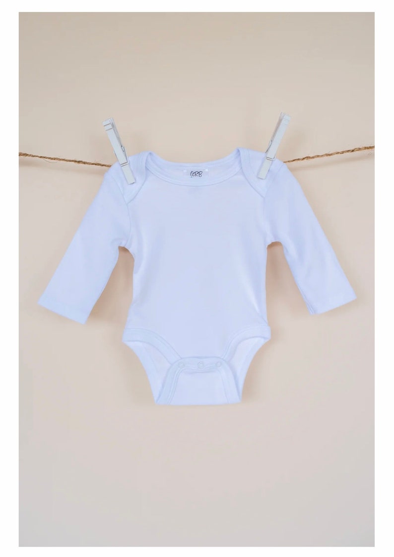 May include: A white long-sleeved baby onesie hanging on a clothesline with two clothespins. The onesie has a small logo on the front that says "6-12M".