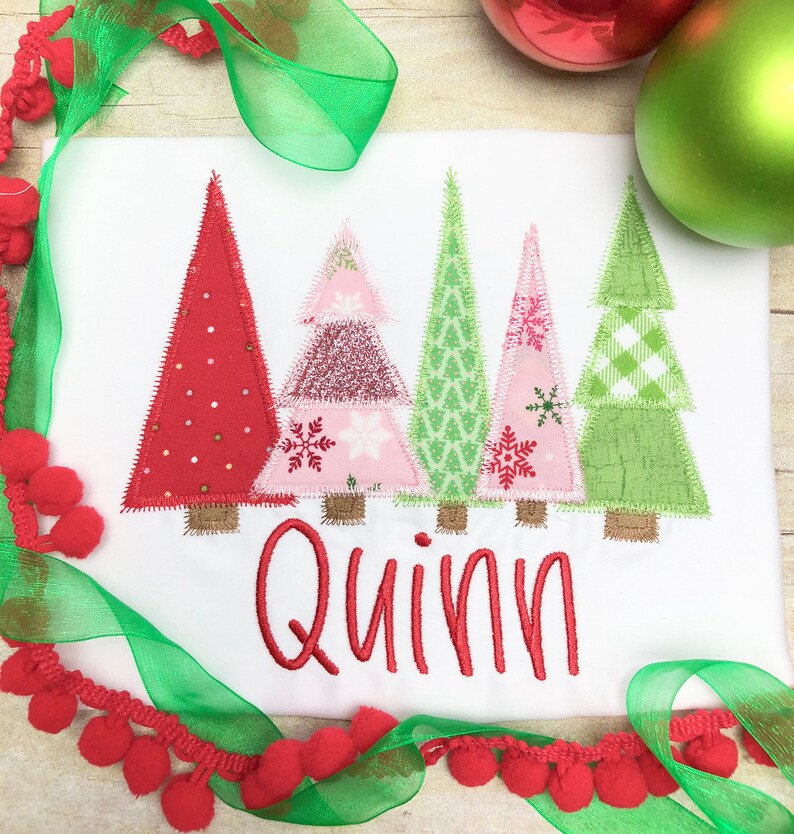 Puede incluir: Un pelele blanco con cinco &aacute;rboles de Navidad de patchwork coloridos y el nombre "Quinn" bordado en rojo.