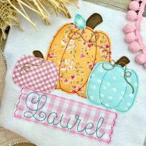 May include: A white onesie with a pink, yellow, and blue applique of three pumpkins. The pumpkins are stitched with a floral pattern and have a gingham pattern on the stems. The onesie has the name "Laurel" embroidered in teal on a pink gingham banner.