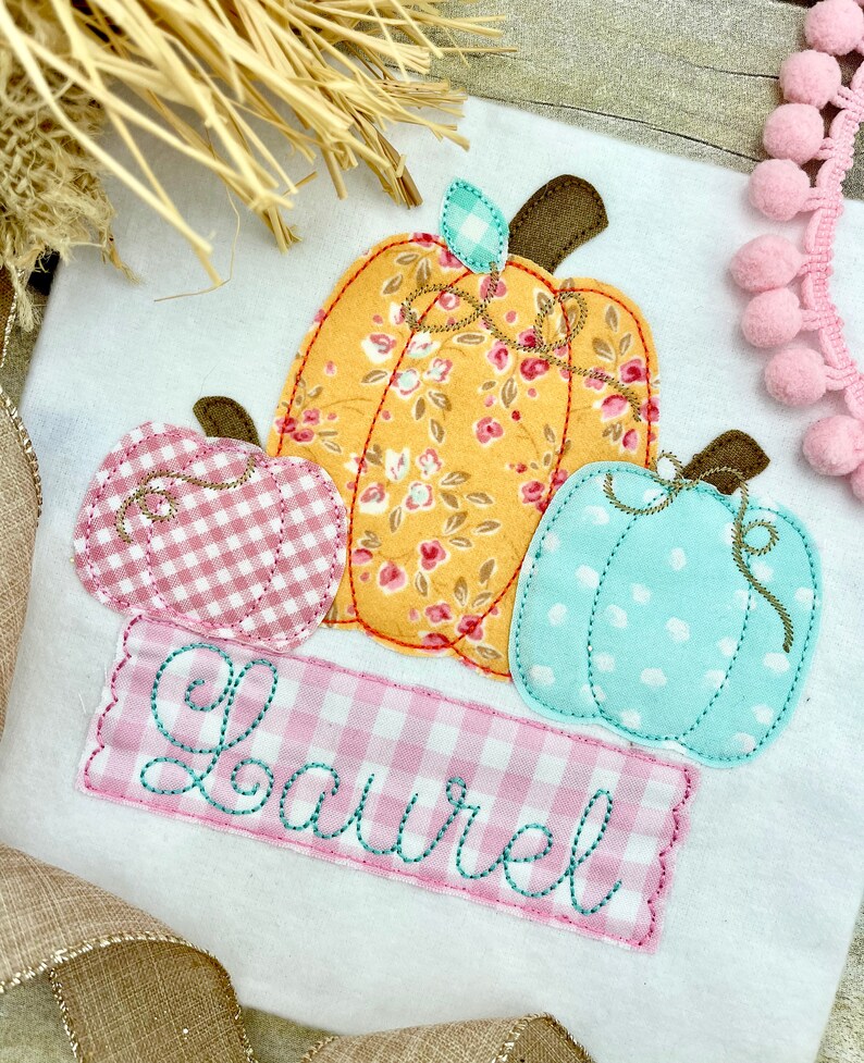 May include: A white onesie with a pink, yellow, and blue applique of three pumpkins. The pumpkins are stitched with a floral pattern and have a gingham pattern on the stems. The onesie has the name "Laurel" embroidered in teal on a pink gingham banner.