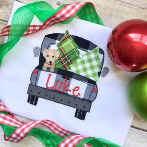 May include: A white t-shirt with an embroidered design of a black and white checkered pickup truck carrying a green and red plaid Christmas tree and a small dog. The name "Luke" is embroidered on the back of the truck.