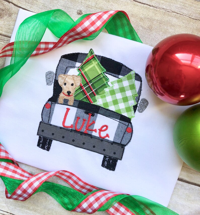 May include: A white t-shirt with an embroidered design of a black and white checkered pickup truck carrying a green and red plaid Christmas tree and a small dog. The name "Luke" is embroidered on the back of the truck.
