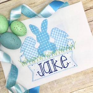 May include: A white embroidered shirt with a blue bunny and two smaller bunnies in a gingham pattern. The bunnies are surrounded by green grass and a banner with the name "Jake" embroidered in blue.