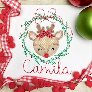 May include: White embroidered Christmas shirt with a red and green reindeer wearing a red bow in a wreath. The name "Camila" is embroidered below the reindeer.