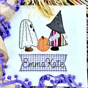 May include: A white fabric panel with an embroidered design of a ghost, a witch, and a pumpkin. The witch is wearing a black hat and a colorful striped dress. The panel has a blue and white gingham banner with the name "Emma Kate" embroidered on it.